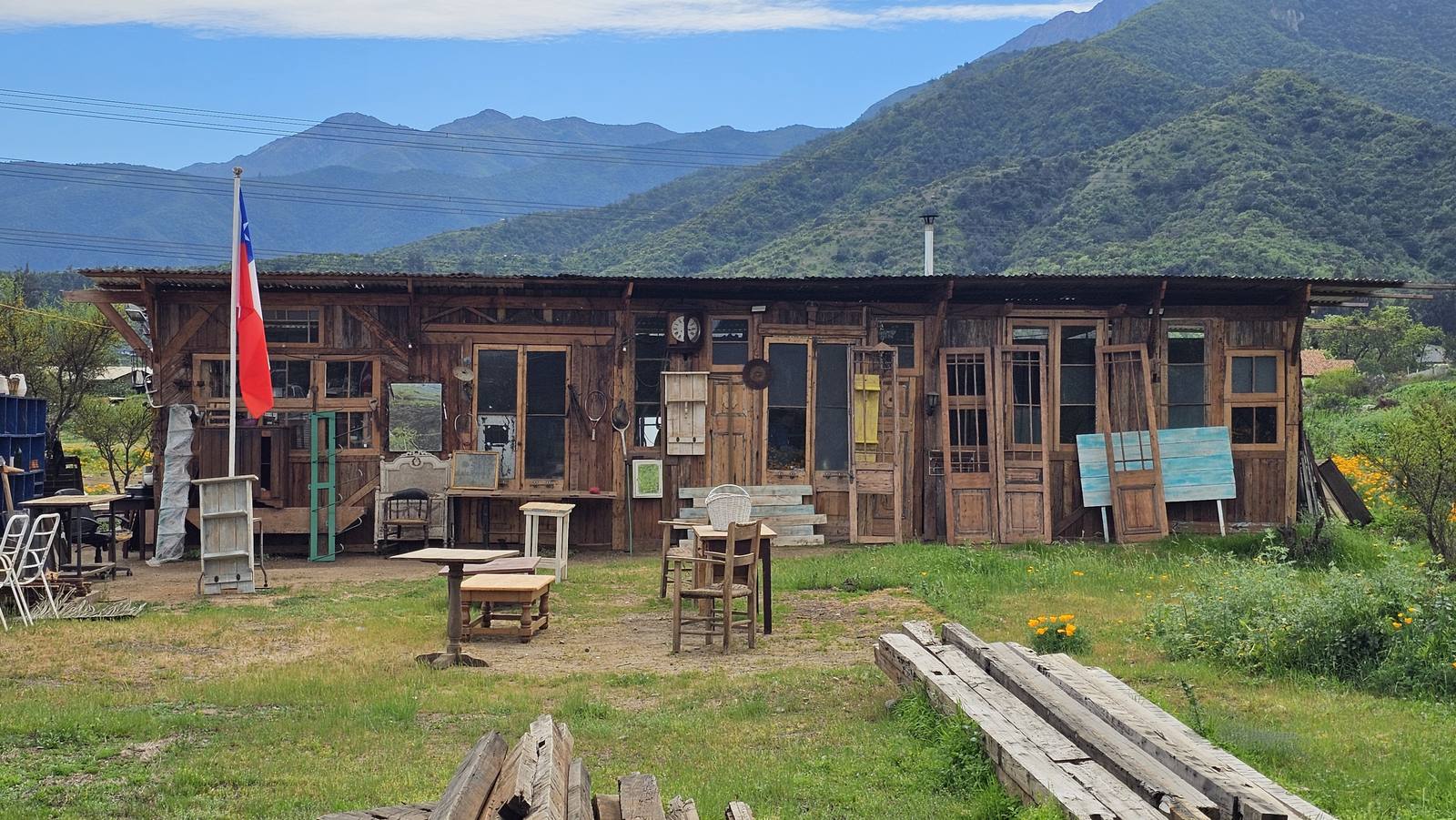El galpón de Antigüedades Roble visto desde afuera: fachada de madera con puertas y ventanas antiguas, tablones apilados en el pasto y el cerro de fondo